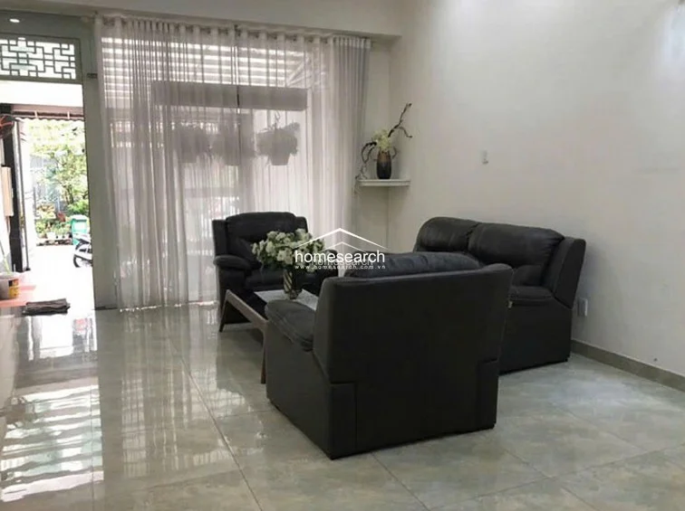 This living room features a modern, open-plan layout with polished light-colored tile flooring that reflects natural light from the large floor-to-ceiling windows. The space is furnished with a dark leather sofa set and connects directly to an outdoor entryway area, creating a seamless flow between indoor and outdoor living.