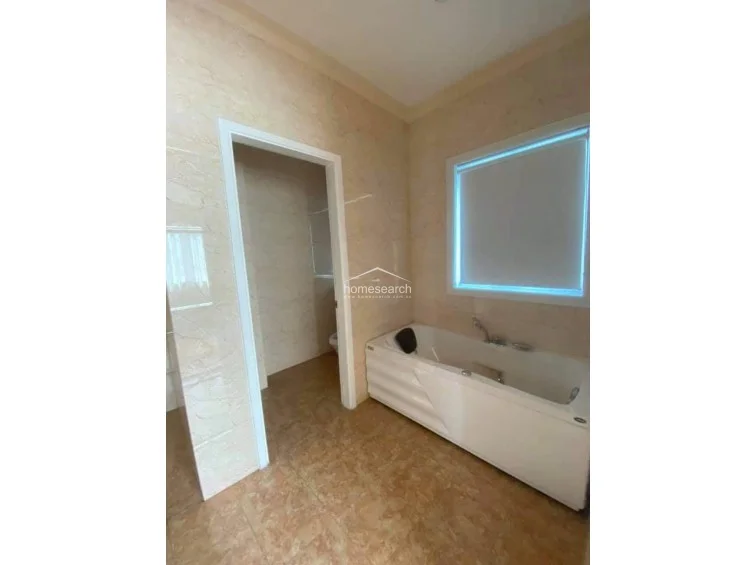 This section of the bathroom highlights a built-in soaking tub positioned beneath a large window that provides natural light, complemented by a roller blind for privacy. The room maintains a cohesive aesthetic with matching marble-style wall tiles and a doorway leading to a separate water closet area, indicating a spacious and functional layout.