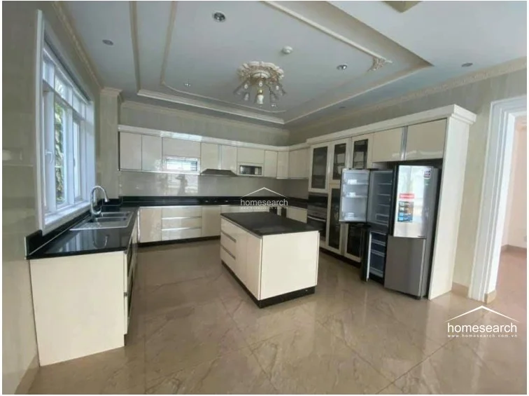 This gourmet kitchen is fully equipped with high-end cabinetry, a central island with a dark stone countertop, and integrated appliances including a large stainless steel refrigerator. The space is well-lit by a combination of natural window light and a decorative chandelier, with polished marble floors that extend throughout the open-concept layout.