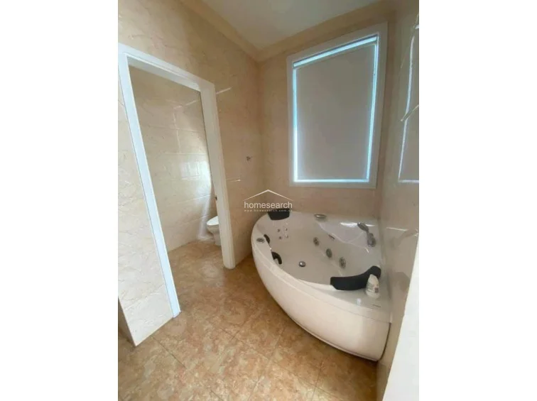 This bathroom is equipped with a large corner jetted tub positioned under a window, offering a spa-like feel with light-toned marble-style wall and floor tiles. A separate, partially visible area contains the toilet, maintaining a functional and private layout.
