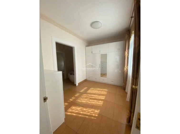 This bedroom features a neutral color scheme with light-colored walls and wood-look tile flooring. It includes a large, white built-in wardrobe with a central mirror and connects directly to an adjacent bathroom, which appears to house a soaking tub.