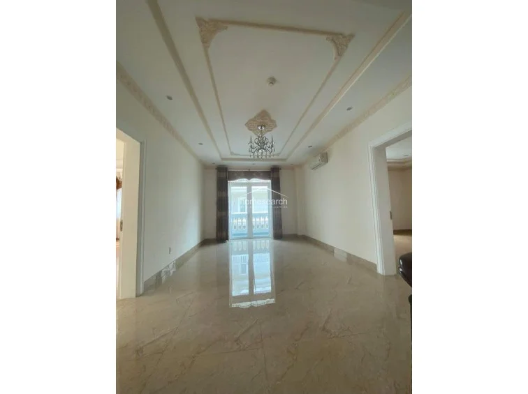 This elegant room serves as a formal living or lounge area, defined by its sophisticated coffered ceiling, intricate crown molding, and a decorative crystal chandelier. The neutral color scheme and glossy marble floors create a bright, airy atmosphere, complemented by a wall-mounted AC unit and classic French doors leading to a balcony.