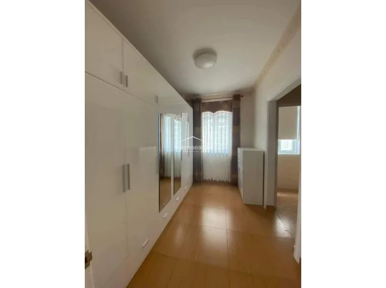 This view highlights the bedroom's layout, emphasizing the extensive white cabinetry that provides ample storage space. The room is illuminated by natural light from a window dressed with floor-length curtains, and the flooring remains consistent with the rest of the suite.