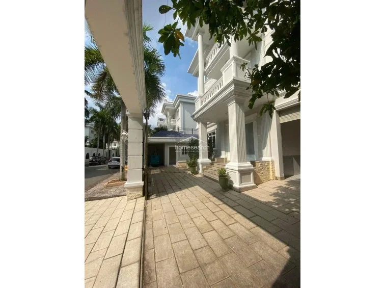 This image displays the exterior driveway and side entrance of a grand, multi-story neoclassical villa featuring prominent white columns and decorative molding. The area is paved with light-colored stone tiles and includes a covered parking section, showcasing a well-maintained, sophisticated architectural style.