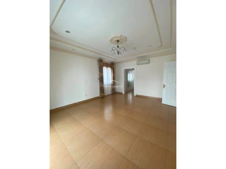 This image shows the open floor plan of the living or bedroom area, which flows seamlessly into an adjoining space. The room features high ceilings with ornate crown molding, light-toned tile floors, and a wall-mounted AC unit, maintaining a bright and well-maintained appearance.