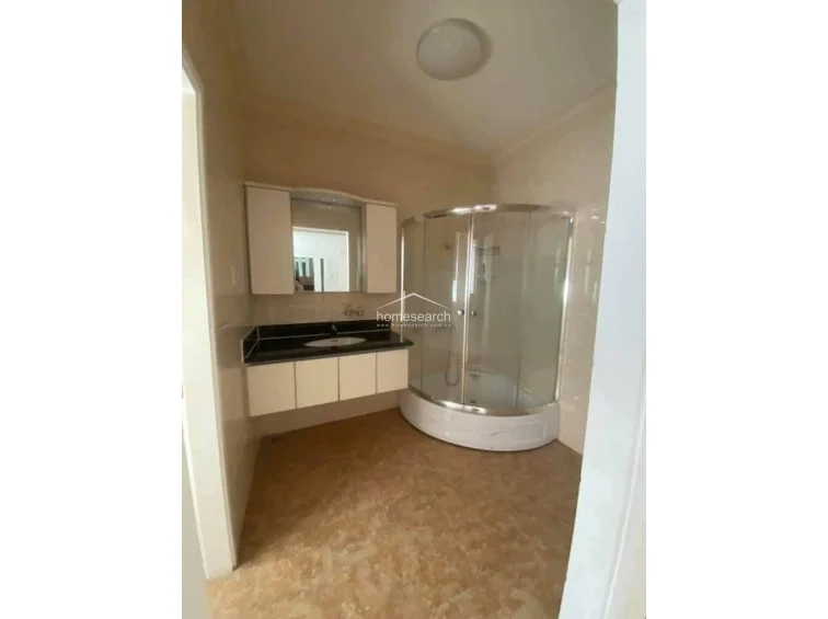 This bathroom features a contemporary floating vanity with a dark stone countertop and a curved glass-enclosed shower stall. The space is finished with light-toned marble-style wall tiles and complementary patterned floor tiles, presenting a clean and well-maintained appearance.