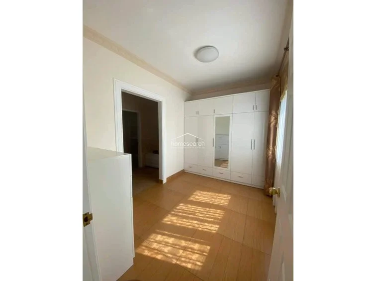 This bedroom features a large, floor-to-ceiling white built-in wardrobe with a central mirror, providing extensive storage. The room is finished with light-colored walls and wood-look tile flooring, creating a clean and neutral aesthetic that connects to an adjacent room through an open doorway.