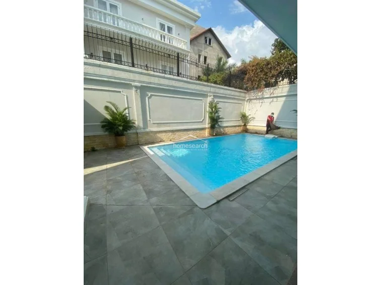 This perspective shows a maintenance worker cleaning the pool, illustrating the ongoing care and upkeep of the property's amenities. The scene captures the contrast between the bright, inviting pool water and the neutral-toned, neoclassical architecture of the home.