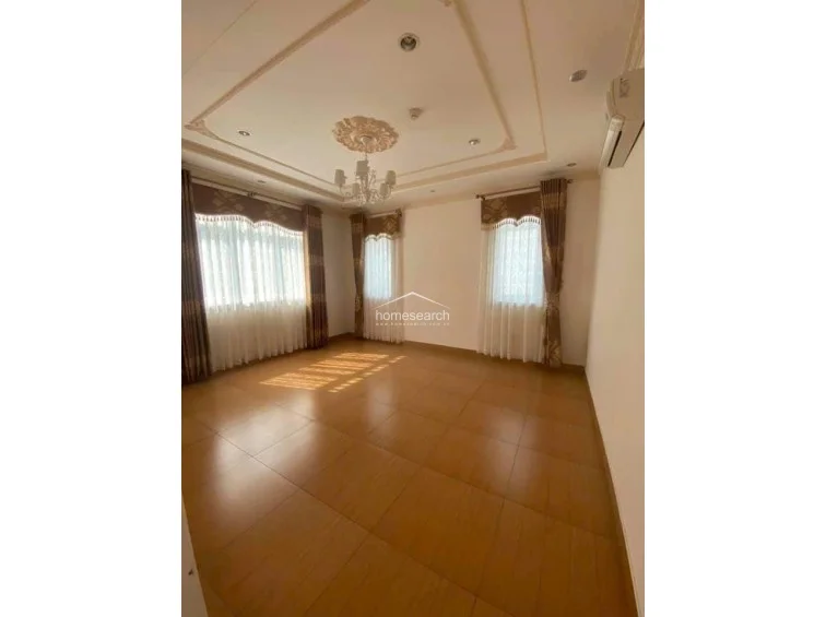 This spacious room serves as a living or primary bedroom area, characterized by elegant crown molding and a decorative ceiling medallion with a chandelier. It is well-lit by multiple windows draped in classic curtains and features a wall-mounted AC unit, with light-toned tile flooring throughout.