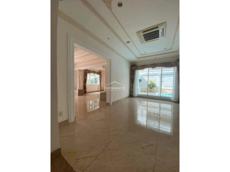 This spacious living area features polished marble flooring and an open-concept flow that connects directly to an adjacent room through a grand, wide doorway. The space is brightened by large glass sliding doors that offer a view of an outdoor pool area, while a recessed ceiling with a central AC unit adds a modern, clean aesthetic.