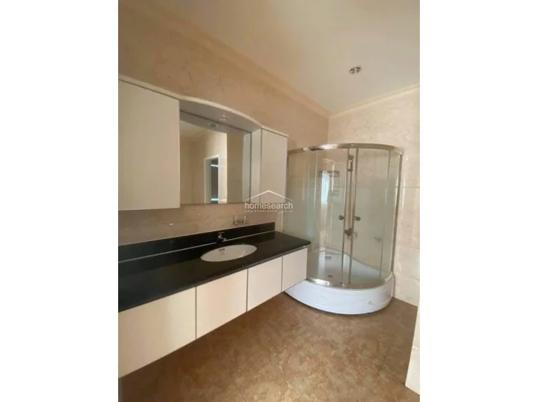 This modern bathroom features a sleek vanity with a black countertop and white cabinetry, paired with a curved glass shower enclosure. The space is finished with warm-toned marble-effect tiles on both the walls and floors, presenting a well-maintained and contemporary appearance.