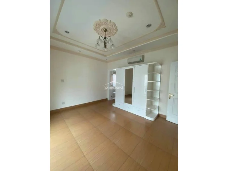 This spacious room is defined by decorative ceiling molding and a classic chandelier, creating an elegant atmosphere. It is equipped with a wall-mounted air conditioning unit and a large white wardrobe with integrated shelving, all set against a clean, neutral backdrop.