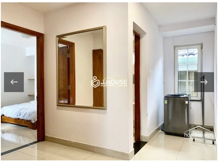 This transition area serves as a functional hallway, connecting the bedroom to a utility space where a washing machine is conveniently located. The space features white walls, tile flooring, and a large wall-mounted mirror, reflecting a practical and efficient use of the property's square footage.