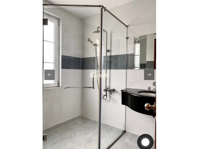 This bathroom features a modern glass-enclosed walk-in shower with a dual-head fixture, set against a backdrop of white wall tiles accented by a dark marble horizontal stripe. The space includes a sleek black vanity countertop and a window that allows for natural light, contributing to a clean and well-maintained aesthetic.