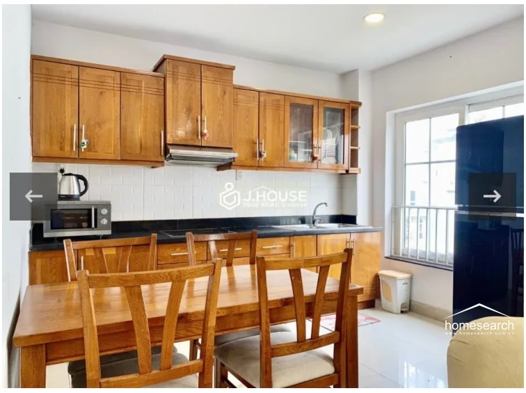 The kitchen area is integrated into the living space, featuring warm-toned wooden cabinetry, a dark countertop, and a white subway-tiled backsplash. It is equipped with essential appliances, including a microwave, electric kettle, and a large refrigerator, all arranged in a functional layout that flows directly into the dining zone.