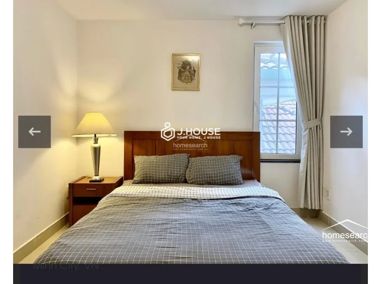 This bedroom features a cozy, minimalist layout with a queen-sized bed framed by a warm-toned wooden headboard and a matching nightstand. The space is finished with neutral white walls and light-colored tile flooring, complemented by a single window draped in floor-length curtains that allows for soft natural light.