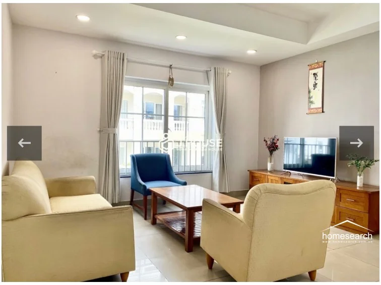 The living room is furnished with a comfortable cream-colored sofa, a matching armchair, and a wooden coffee table, providing a cozy seating arrangement. A wooden media console sits against the wall, and the room is accented with tasteful decor like a wall hanging and a small floral arrangement.