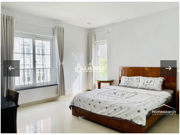 This bright bedroom offers a functional layout with a large window and a smaller side door providing ample natural light and views of the surrounding urban architecture. The room is furnished with a wooden bed frame, a dedicated desk area, and a vanity, all set against a clean, white-walled backdrop with polished tile floors.