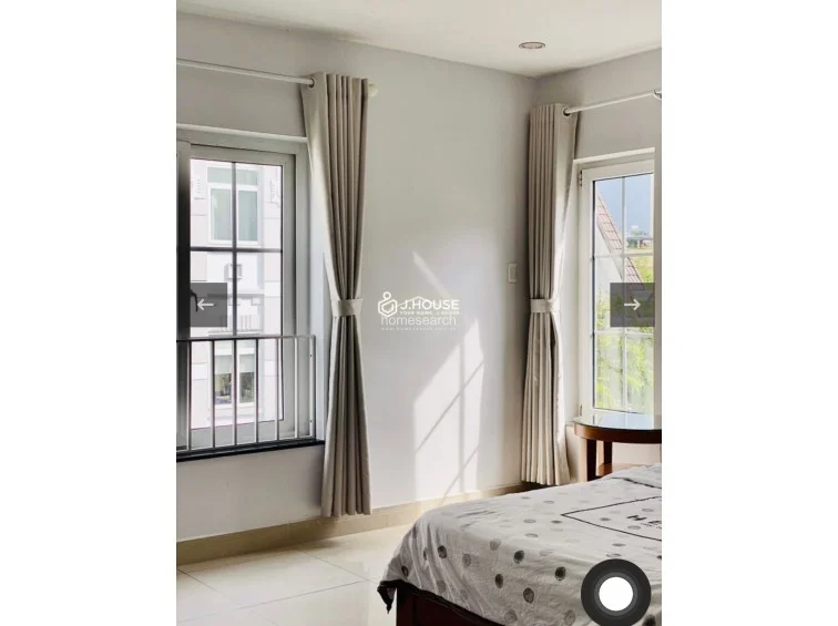 This image captures the corner of the bedroom where two windows meet, allowing for cross-ventilation and significant natural light. The simple, modern furniture and neutral color palette emphasize a clean, well-maintained living environment.