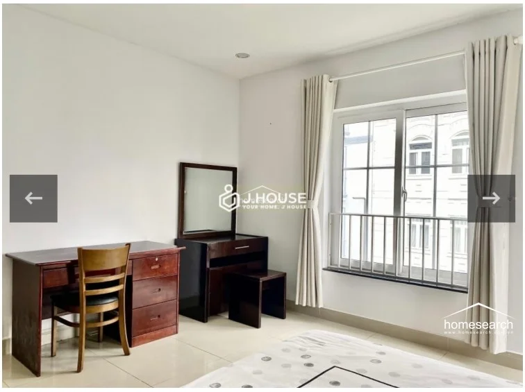 This view highlights the bedroom's workspace, featuring a sturdy wooden desk and a matching vanity with an integrated mirror. The area is positioned near a window with floor-length curtains, creating a well-lit, practical corner that maintains the room's cohesive, wood-accented aesthetic.