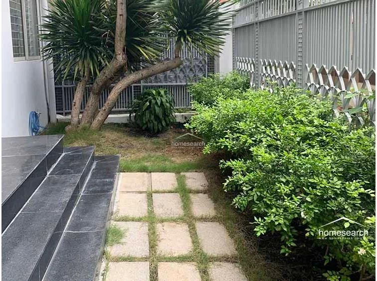 This outdoor area features a small garden space with lush greenery and a mature tree, bordered by a decorative metal fence. The transition to the interior is marked by a set of dark, polished stone steps, suggesting a well-maintained and structured exterior design.