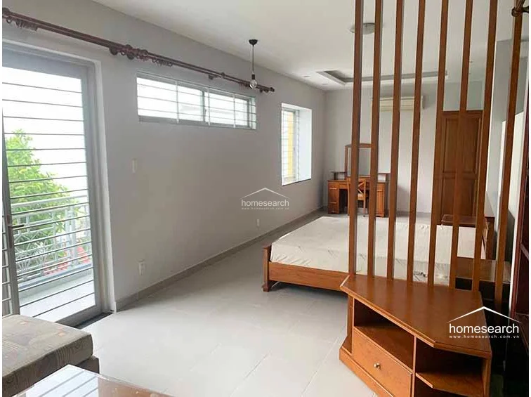 This spacious bedroom is divided by a vertical wooden slat screen, creating a semi-private sleeping area while allowing light to travel from the large balcony door. The room is furnished with a wooden bed frame and vanity, complemented by light-colored tile floors and a neutral color scheme that enhances the sense of space.
