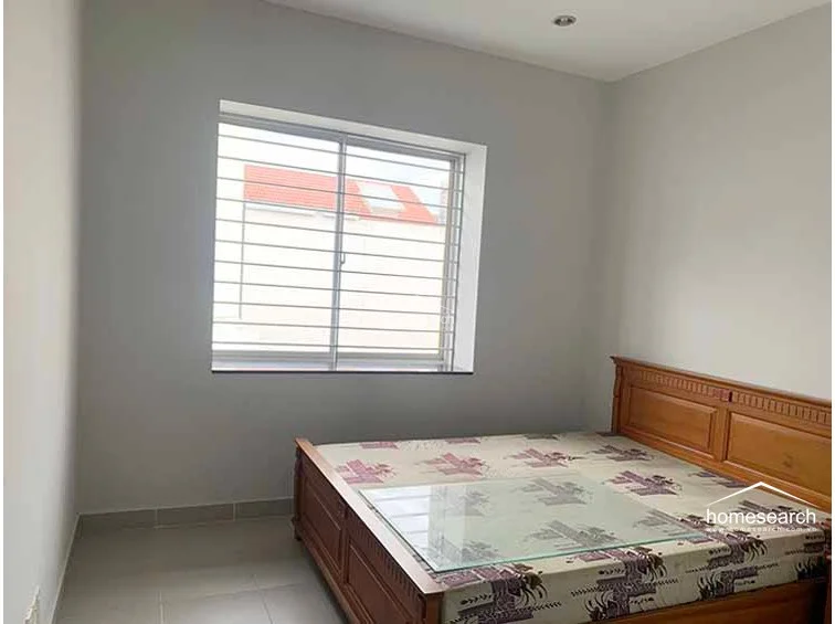 This bedroom is finished with simple, light-toned walls and matching floor tiles, creating a clean and minimalist aesthetic. The room is furnished with a sturdy wooden bed frame and features a large window with security bars that provides natural light and a view of the neighboring rooftops.