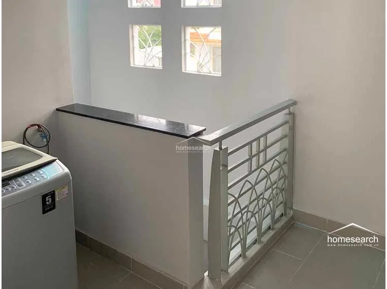 This utility or laundry area is located on an upper landing, featuring a functional layout with a top-loading washing machine. The space is brightened by four square windows with decorative metal grilles and is finished with neutral walls and consistent floor tiling throughout.