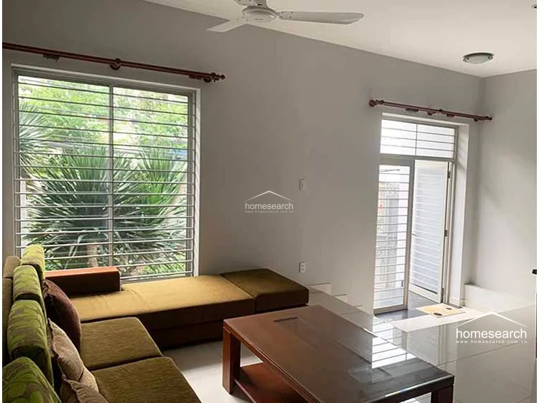 This living area is bright and airy, featuring large windows with security bars that allow for ample natural light. The room is furnished with a modern olive-toned sofa and a dark wood coffee table, set against neutral walls and a ceiling fan for climate control.