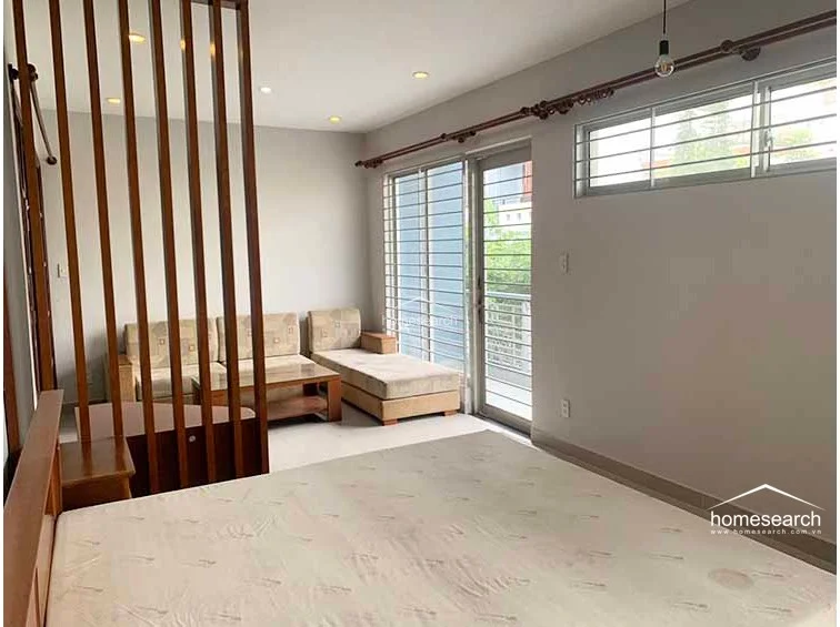This bedroom layout incorporates a minimalist aesthetic with a neutral color palette and clean lines. A wooden room divider adds architectural interest and separates the sleeping area from a cozy lounge corner, which features a sofa and direct access to a balcony through a glass-paneled door.