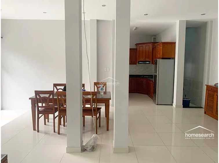 This open-concept dining and kitchen area is defined by large white structural columns that create a distinct flow between the spaces. The kitchen is equipped with warm-toned wooden cabinetry and a stainless steel refrigerator, while the dining area features a matching wooden table set on clean, light-colored tile flooring.