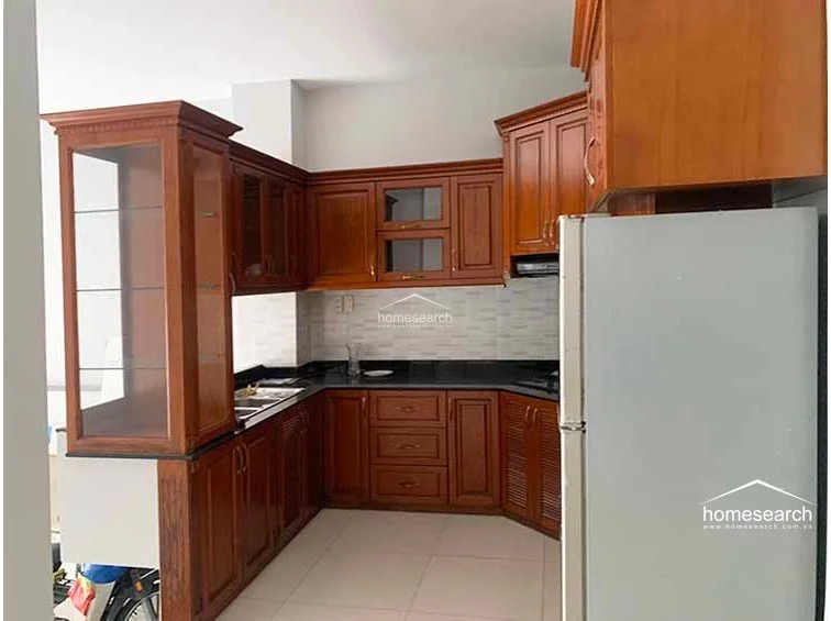 This L-shaped kitchen features warm-toned wooden cabinetry with a mix of solid and glass-fronted doors, providing ample storage and a classic aesthetic. It is equipped with a dark countertop and a light-colored tiled backsplash, maintaining a clean and well-kept appearance.