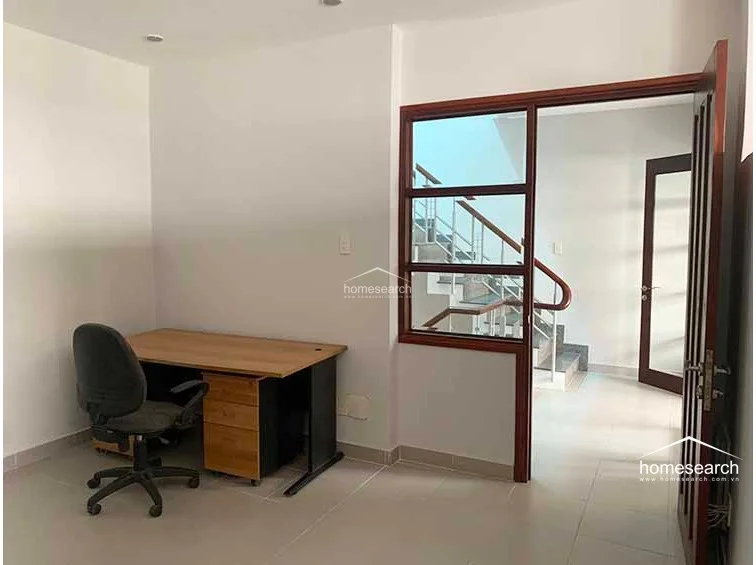 This functional home office or study area features neutral white walls and light-colored floor tiles, creating a bright and minimalist workspace. A large, multi-paned window provides a direct view into the adjacent stairwell, promoting an open flow between the rooms.