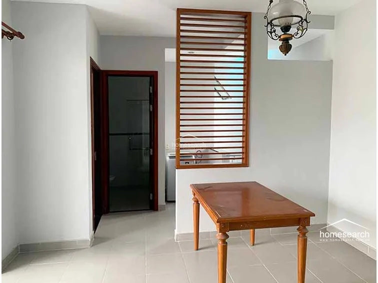 This dining or multipurpose area is defined by a decorative wooden slat partition that adds architectural interest while maintaining an open-concept feel. The space is illuminated by a classic pendant light fixture and includes a simple wooden table, set against a backdrop of light gray walls and tiled flooring.
