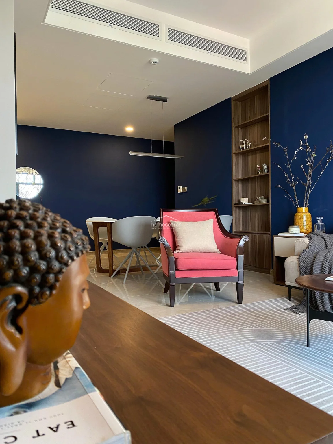 This intimate perspective focuses on the transition between the living and dining areas, framed by a decorative Buddha sculpture in the foreground. The deep blue wall continues into the dining space, where a tall, built-in wooden shelving unit provides elegant display storage. The layout is well-planned, creating a harmonious balance between the comfortable lounge seating and the formal dining area.