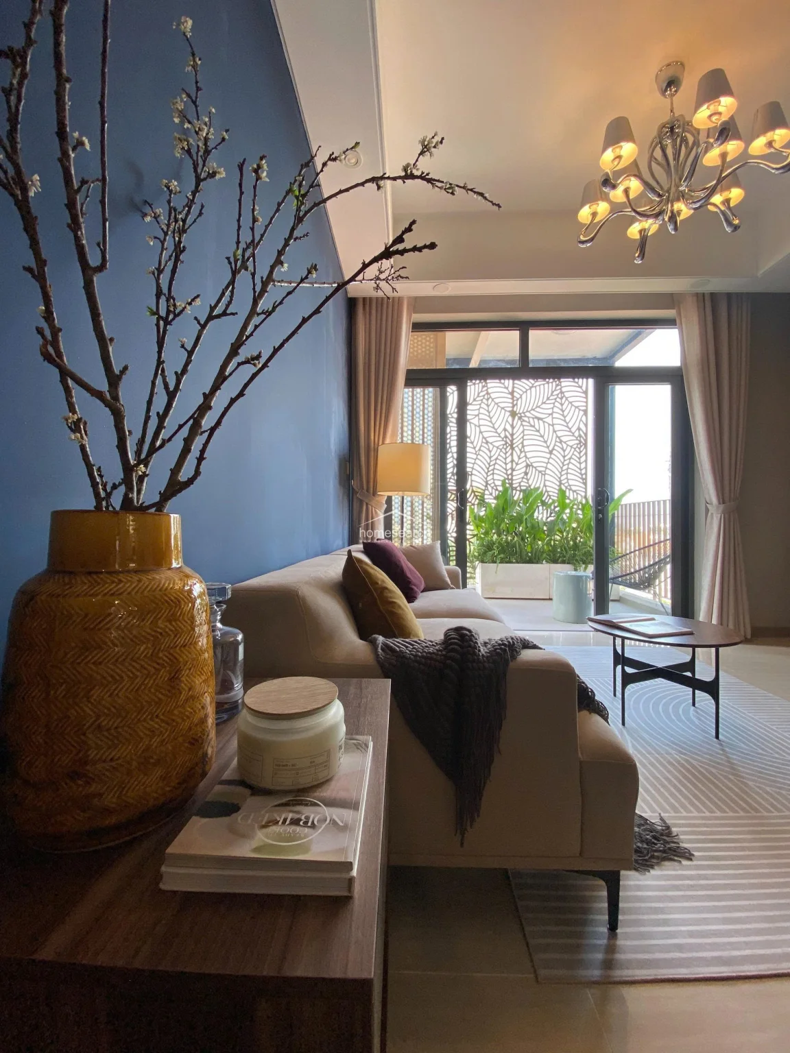 This image showcases a cozy living area featuring a bold navy accent wall and a cream-colored sofa, creating a sophisticated contrast. The space flows seamlessly toward a balcony through large glass doors, which allow for ample natural light and a view of lush greenery. The room is finished with light-colored tile flooring, a decorative chandelier, and a built-in wooden bookshelf that adds warmth to the modern design.