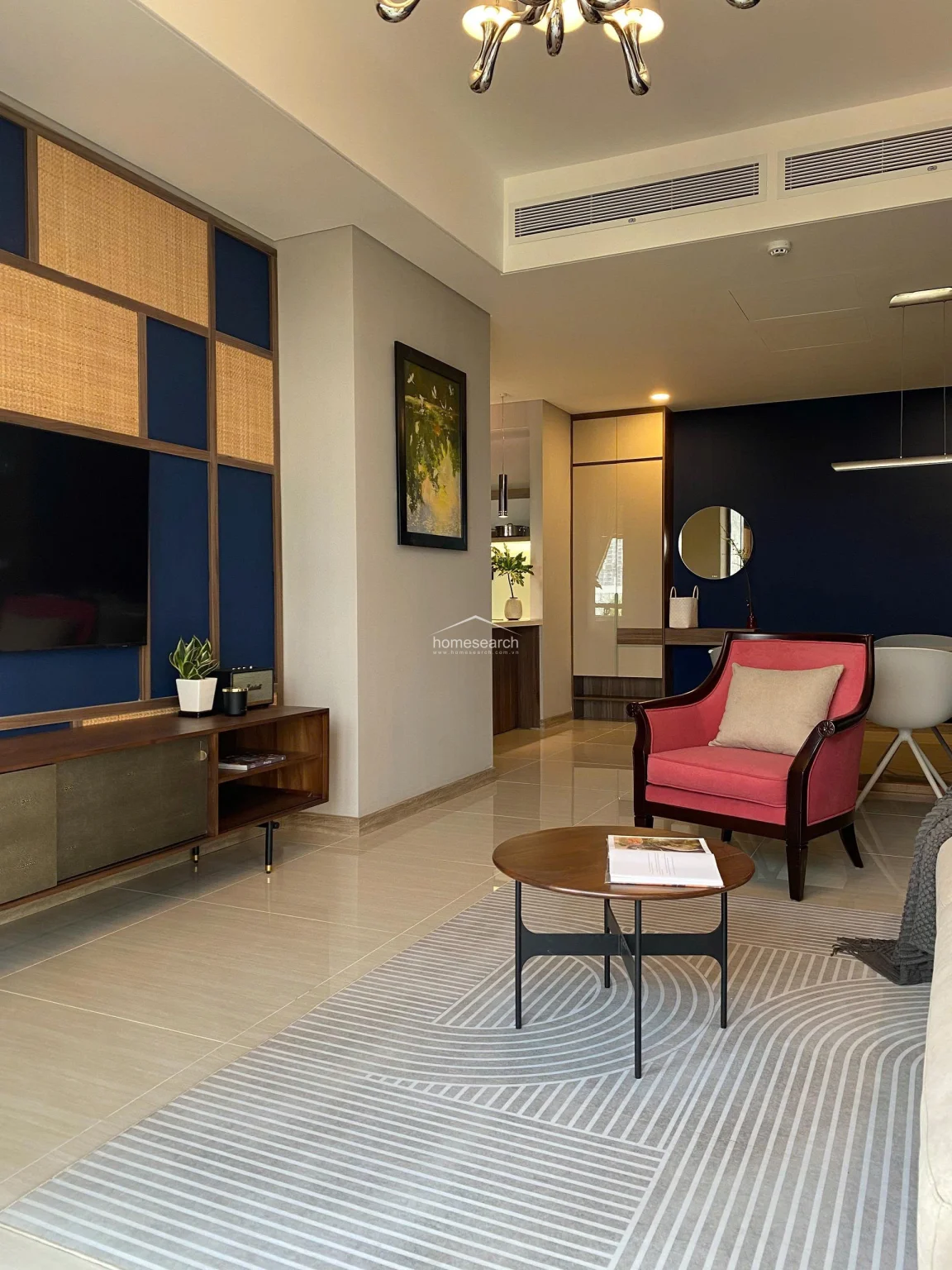 The living room showcases a custom media wall with rattan and navy blue paneling, complemented by a sleek wooden console and a cozy pink armchair. A central ceiling-mounted air conditioning unit ensures climate control, while the neutral color palette and minimalist furniture maintain a clean, high-end aesthetic.