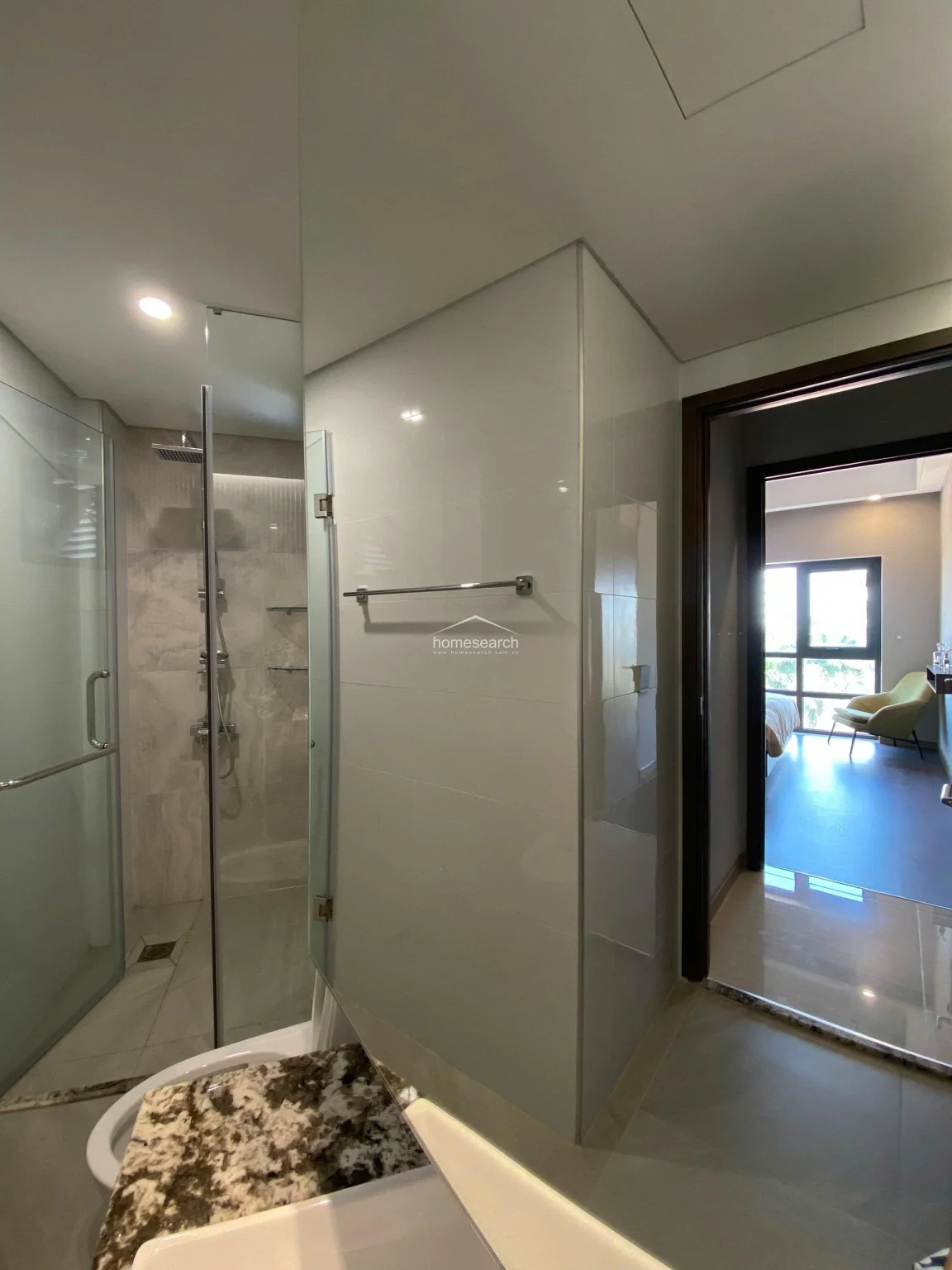 This perspective looks from the bathroom back into the bedroom, emphasizing the open-concept layout and the use of glass partitioning to enhance natural light flow. The transition between the tiled bathroom floor and the bedroom's wood-look flooring is clean and well-executed. The room maintains a cohesive, modern design language with neutral tones and high-quality finishes throughout.