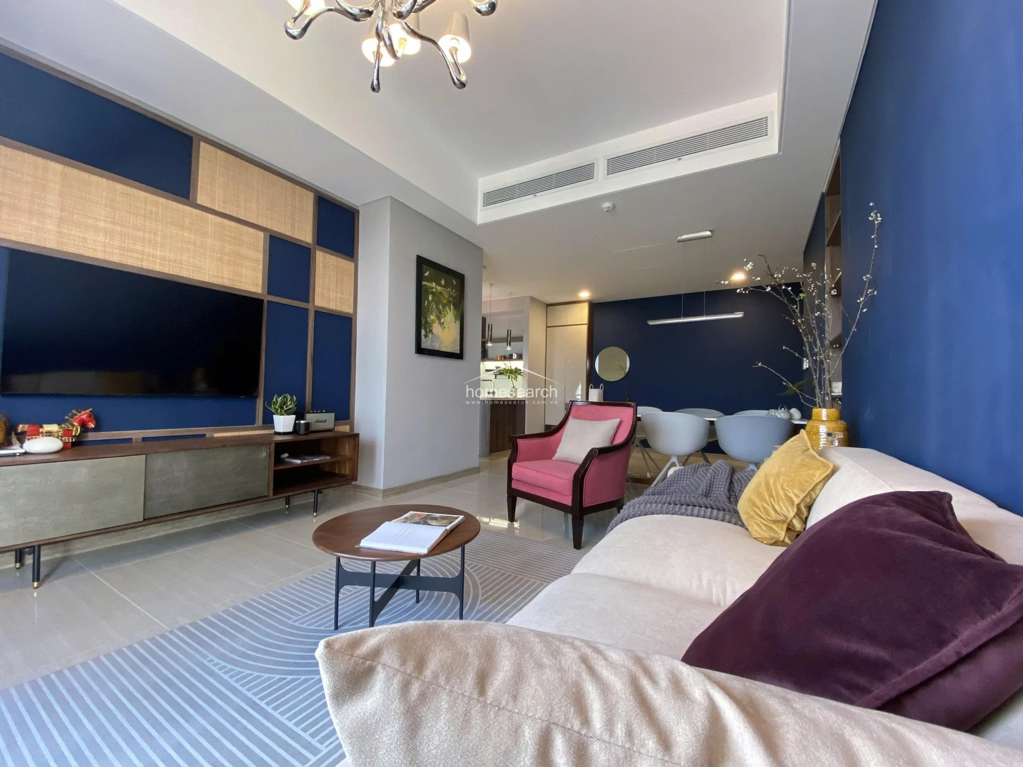 The open-concept living room is defined by a striking deep blue accent wall and a sophisticated media console with integrated cane-webbing details. The space is brightened by recessed ceiling lighting and a modern chandelier, while the neutral-toned sofa and pink armchair create a cozy, well-curated seating arrangement.