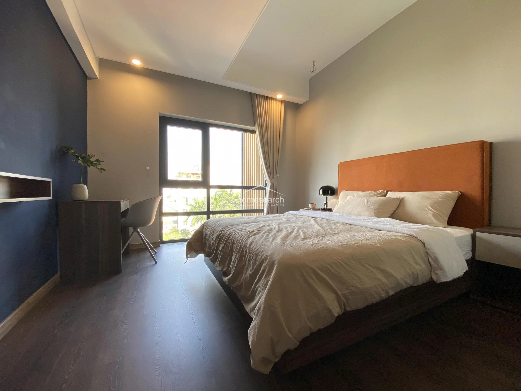 This modern bedroom features a striking contrast between a deep navy accent wall and soft grey tones, complemented by dark wood-look flooring. The room is anchored by a large bed with a vibrant orange headboard and benefits from natural light streaming through a floor-to-ceiling window, which also provides a view of the surrounding greenery. A sleek, minimalist desk setup is integrated into the layout, creating a functional workspace within the bedroom.