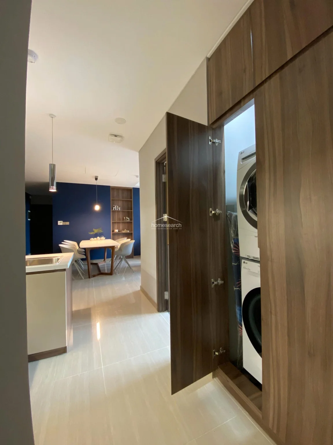 This functional hallway includes a discreet, built-in floor-to-ceiling wooden cabinet that houses a stacked LG washer and dryer unit. The space maintains a clean, minimalist aesthetic with polished light-colored floor tiles and a direct, open sightline into the dining and kitchen area.