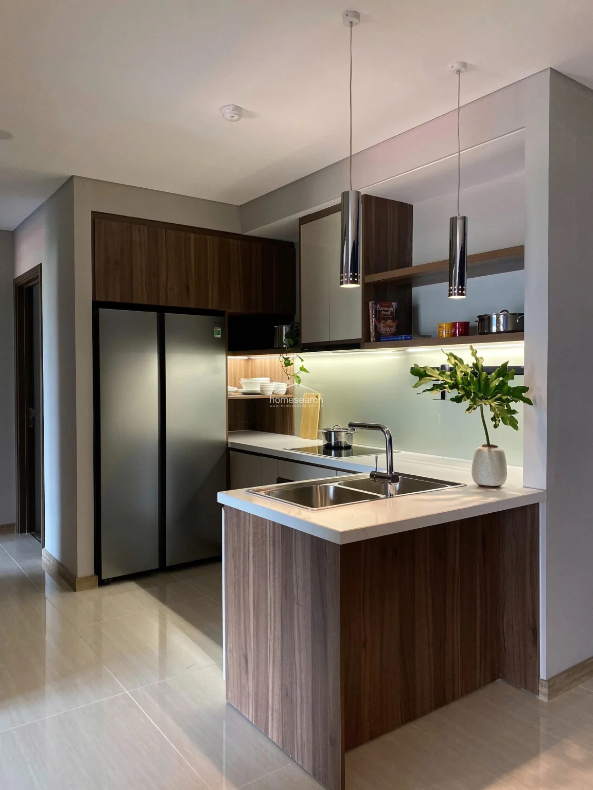 This modern, compact kitchen features high-quality wood-grain cabinetry and a sleek white countertop with an integrated sink. It is equipped with a full-sized stainless steel refrigerator and under-cabinet lighting, all set against a neutral, polished tile floor that flows seamlessly into the adjacent living area.