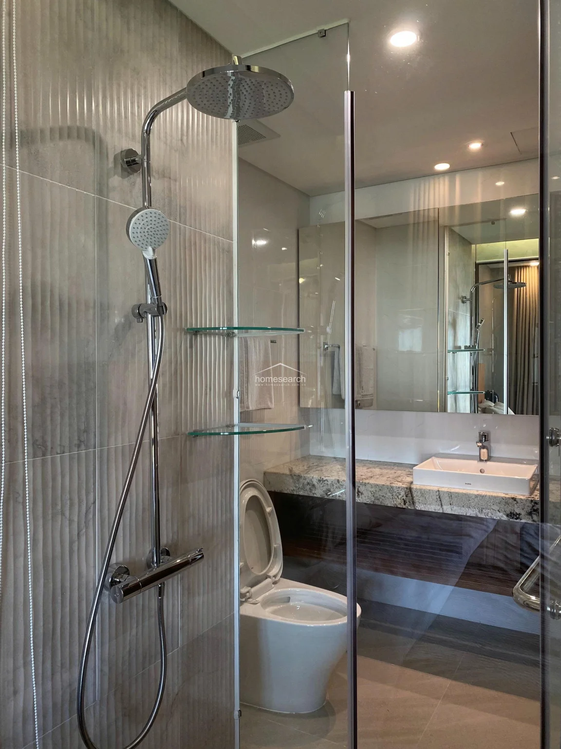 The bathroom features a sophisticated design with a rainfall showerhead and a sleek chrome shower system mounted against textured, light-grey wall tiles. A floating granite-topped vanity provides a luxurious touch, while the glass shower enclosure ensures the space remains bright and spacious. The overall condition is excellent, reflecting a high-end, contemporary aesthetic.
