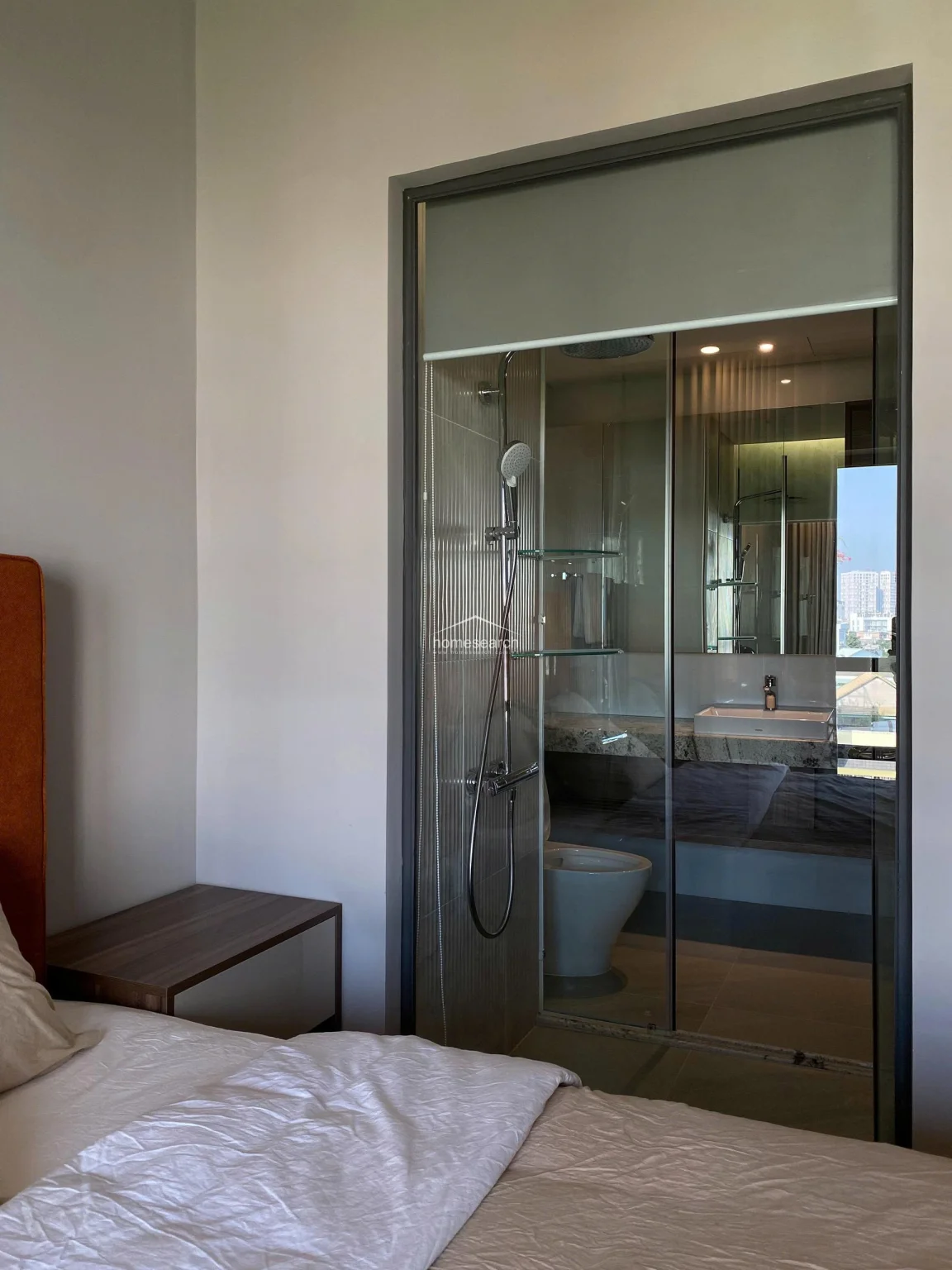 This view highlights the en-suite bathroom, which is uniquely separated from the bedroom by a glass partition, creating an open and airy feel. The bathroom is equipped with modern fixtures, including a wall-mounted shower and a contemporary sink, all set against a neutral, tiled backdrop. The design emphasizes a seamless flow between the sleeping and bathing areas, maintaining a high standard of modern maintenance and style.