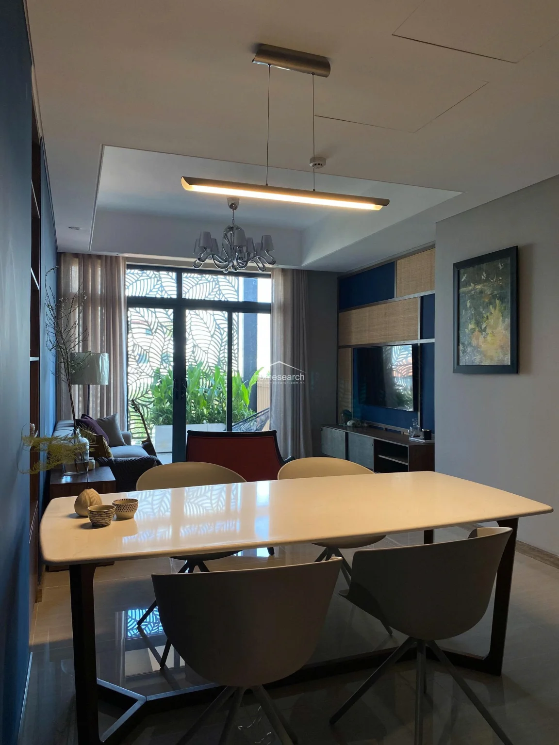 This view highlights the open-concept dining and living area, emphasizing the cohesive color palette of deep blue and neutral tones. The dining space is anchored by a modern white table with contemporary chairs, positioned under a minimalist linear pendant light. The room maintains a high level of maintenance and features integrated ceiling air conditioning units and a stylish media wall with rattan accents.