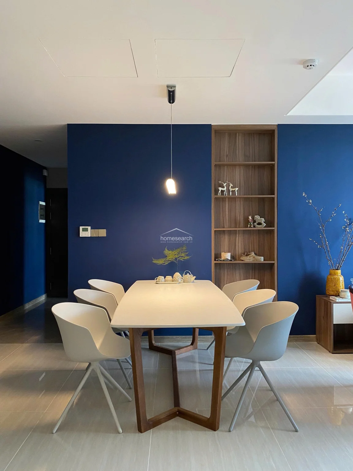 This image provides a clear view of the dining area, which is illuminated by a sleek, singular pendant light that creates a focal point above the table. The space is characterized by clean lines, modern white dining chairs, and a polished tile floor that reflects the light. The integrated wooden shelving and the deep blue wall finish reinforce the sophisticated, well-maintained aesthetic of the home.