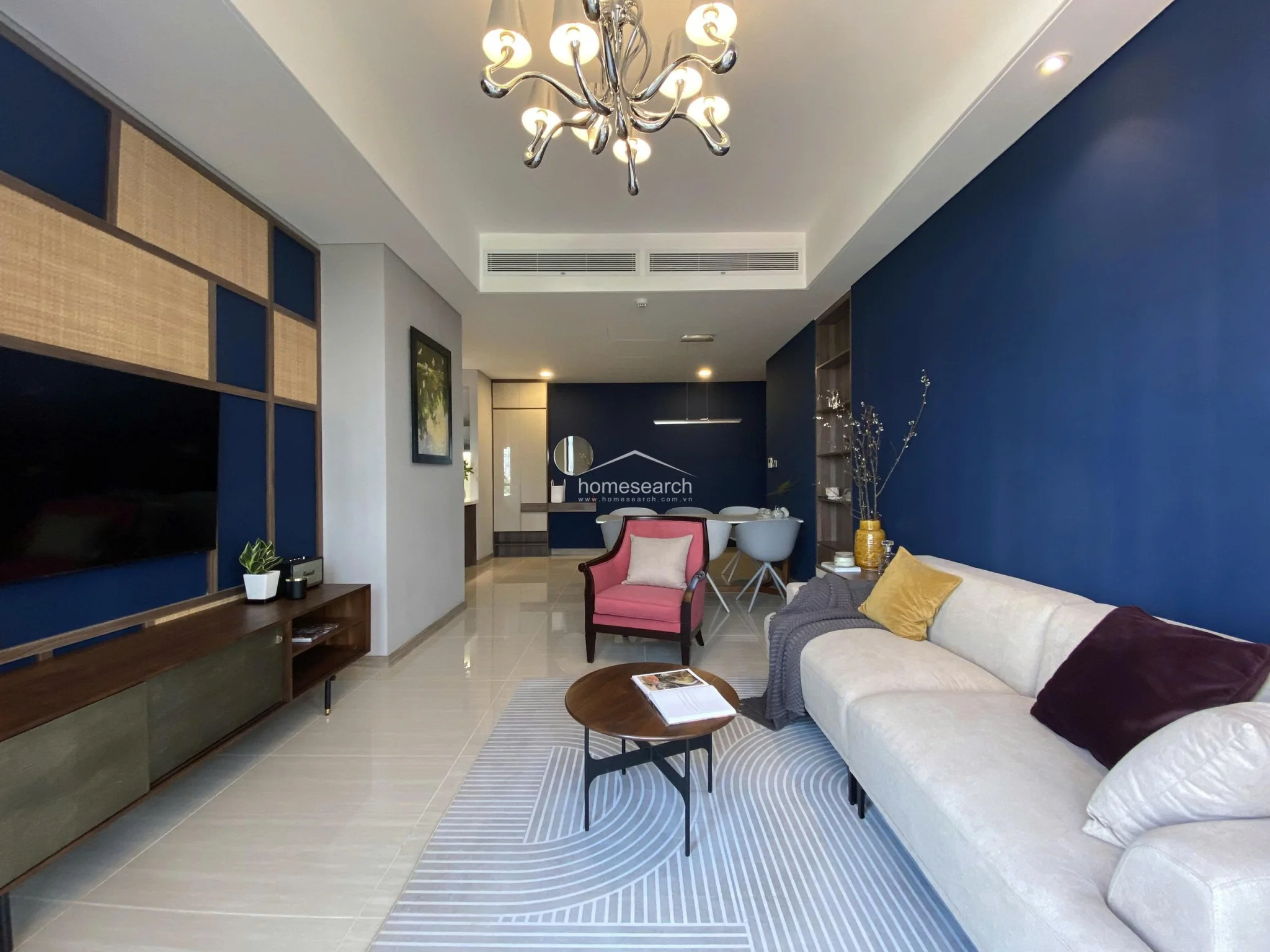 This wide-angle shot captures the entire living and dining flow, highlighting the spaciousness and the intentional use of color to define different zones. A vibrant pink accent chair adds a pop of color to the neutral sofa area, while the deep blue walls provide a dramatic backdrop throughout the entire room. The property appears to be in excellent condition, featuring recessed lighting and high-quality finishes throughout.