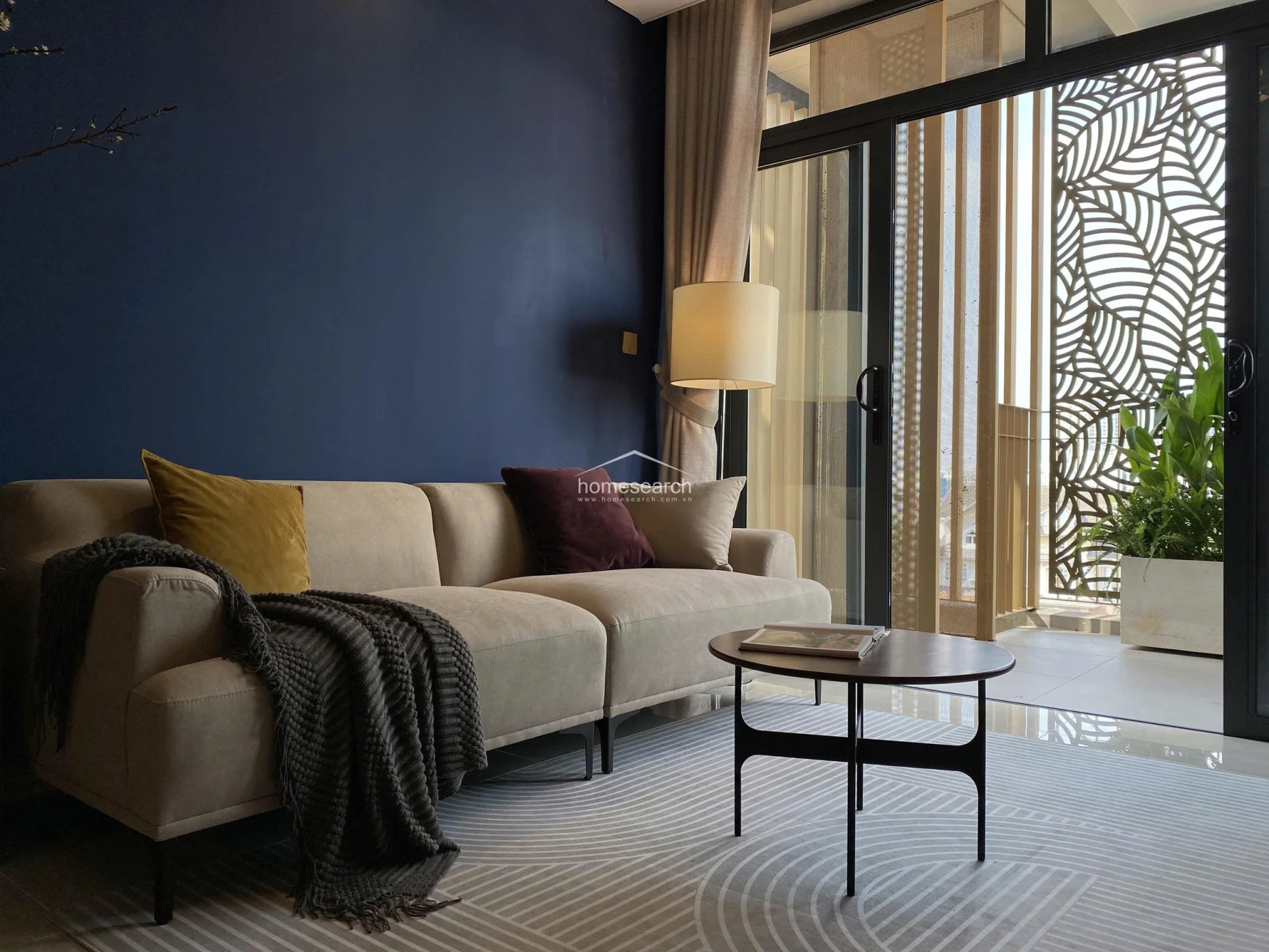This cozy living area centers around a plush beige sofa accented with mustard and plum cushions, positioned next to a floor lamp for soft ambient lighting. Large sliding glass doors lead to a balcony featuring an intricate, leaf-patterned metal screen that provides both privacy and a decorative architectural element.
