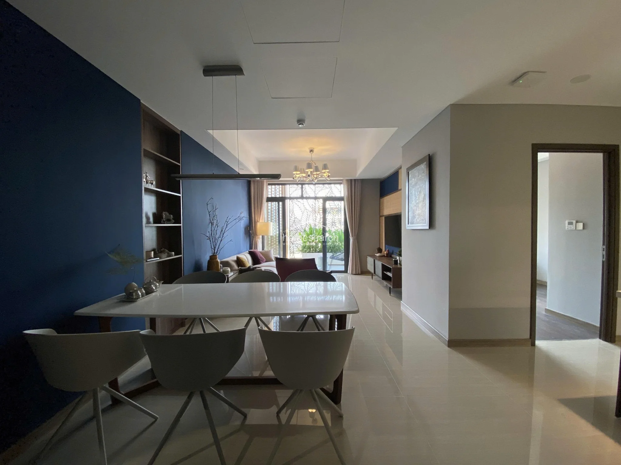 This open-concept living and dining area features a sophisticated navy blue accent wall with integrated shelving and polished light-colored tile flooring. The space flows seamlessly toward a private balcony, which is framed by elegant floor-to-ceiling curtains and illuminated by a modern chandelier.
