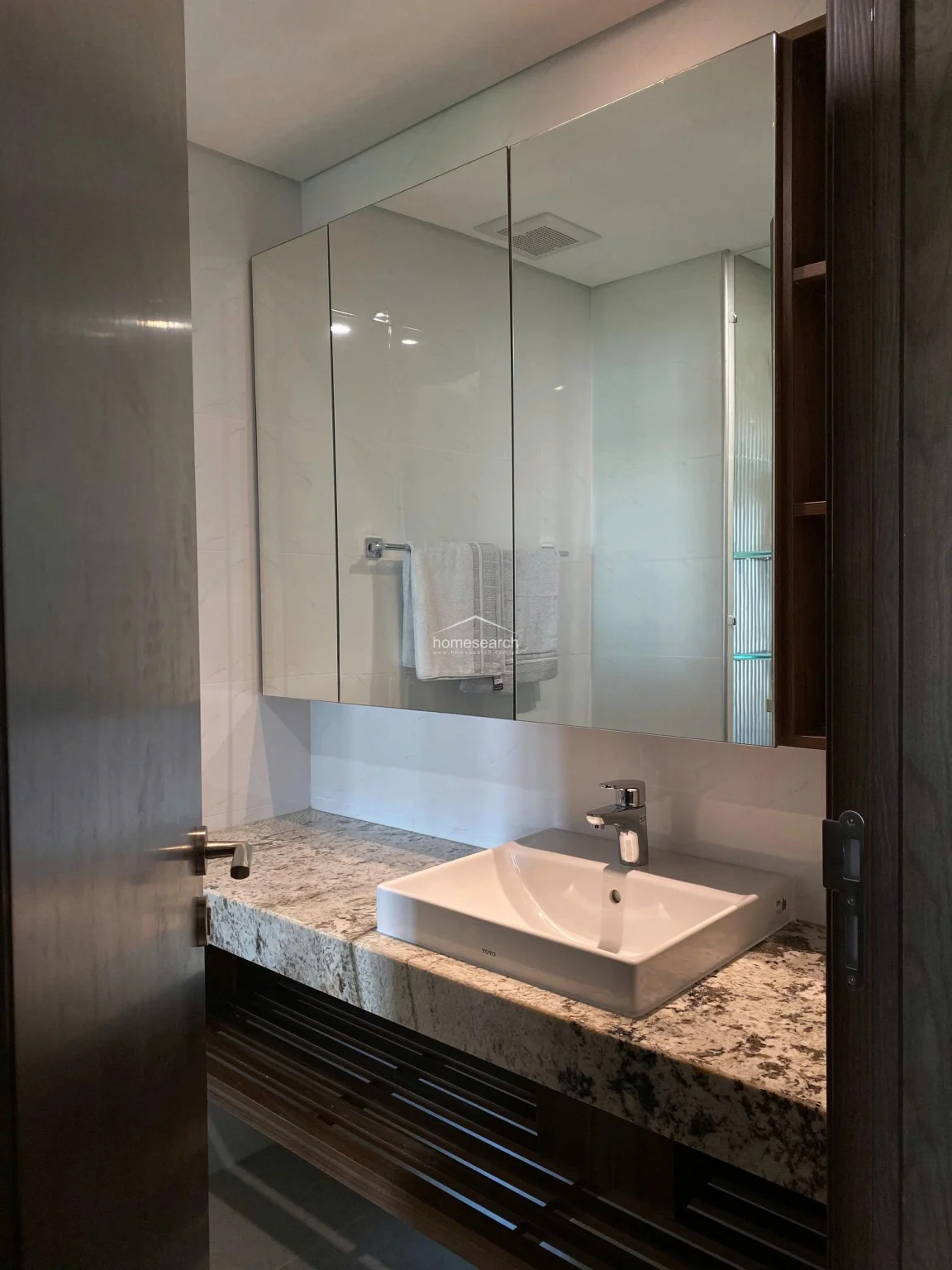 This compact, well-designed bathroom vanity area emphasizes functionality with a large, integrated triple-mirror cabinet that provides ample hidden storage. The space is anchored by a premium TOTO basin and a sophisticated granite countertop, complemented by a dark wood slatted base for additional storage. The neutral wall tiles and warm wood accents create a polished, high-maintenance look that is both practical and elegant.
